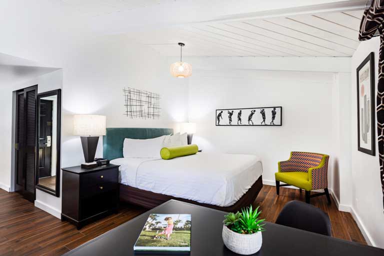 hotel room bed with white sheets, green retro pillow, white walls, and bright natural light