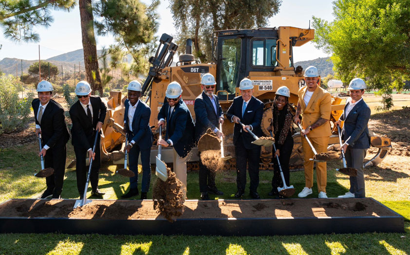 9 people standing in suites with white construction hard hats and shovels digging hole in a garden box while smiling.