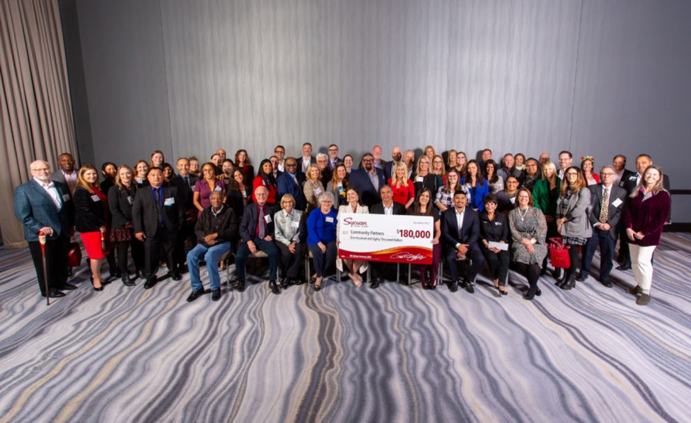 Large crowd of smiling people that's sitting down in the front and also standing in the back are holding a check that says it's from Sycuan Casino that's written out for $180,000